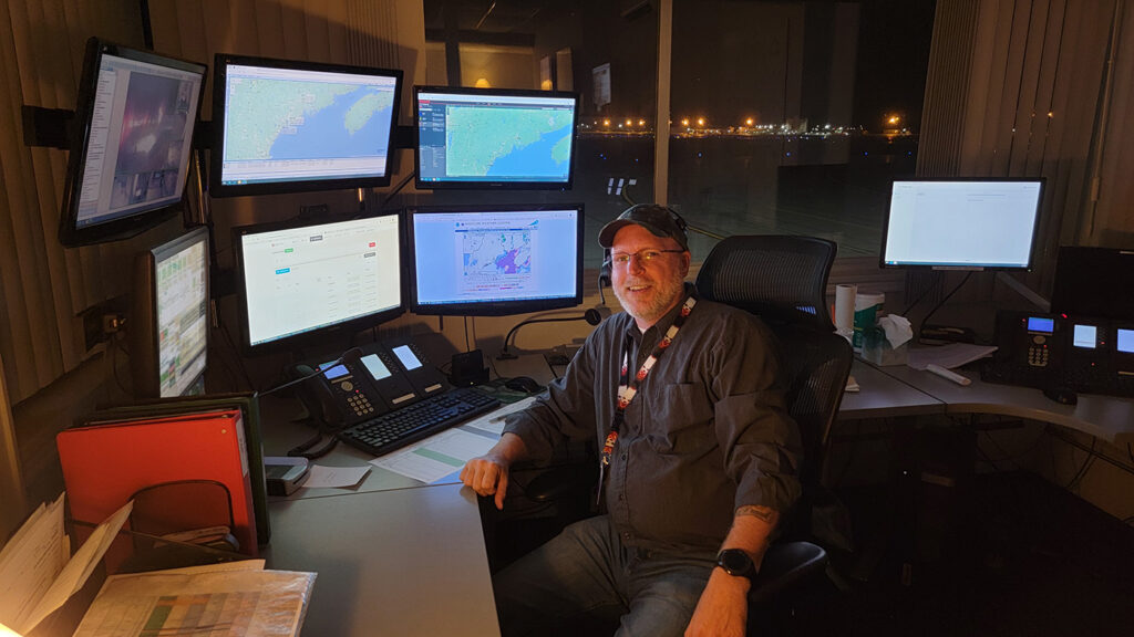 LifeFlight of Maine Communications Supervisor Jeremy Bean sitting at his desk, surrounded by six monitors showing maps and landing zone information