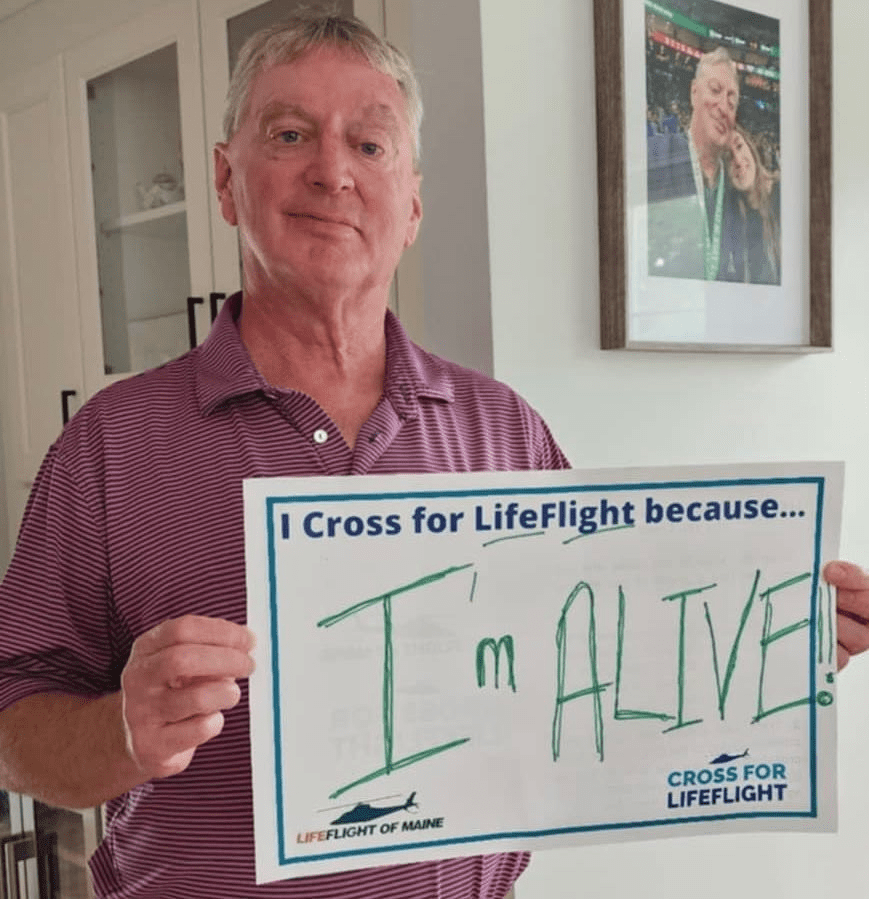 John holds a sign saying "I Cross for LifeFlight because... I'm Alive," as part of the 2025 Cross for LifeFlight.